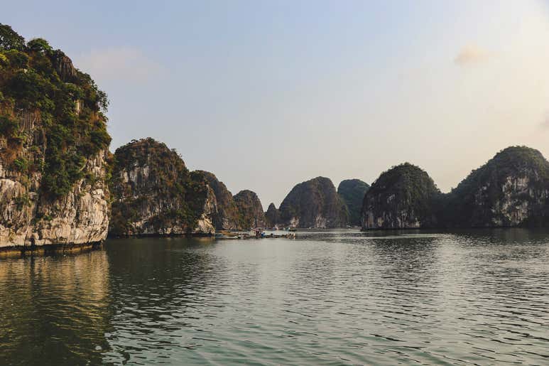 Green-covered limestone karsts of Ha Long Bay stand against a soft sky, and small boats navigating the peaceful waters below.