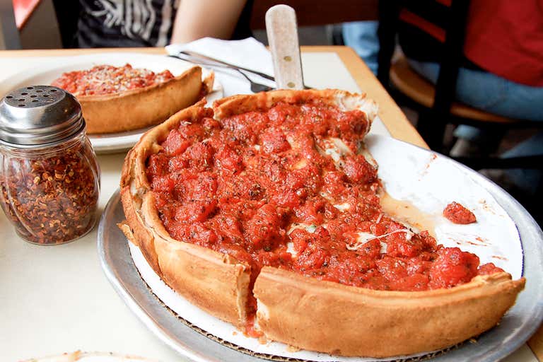 Una pizza estilo deep-dish servida en la mesa de un restaurante con un frasco de hojuelas de chile rojo.