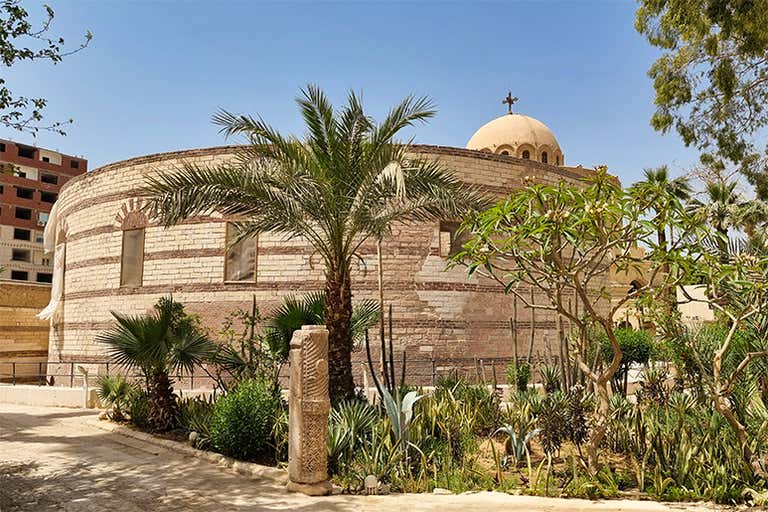 Circular brick church surrounded by lush greenery, palm trees, and ancient stone carvings under a clear blue sky.
