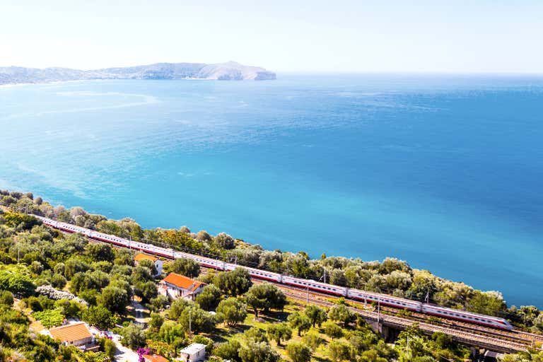 Ein malerischer Blick auf die italienische Küste mit einem Zug, der durch üppiges Grün fährt, während das Meer im Hintergrund zu sehen ist.