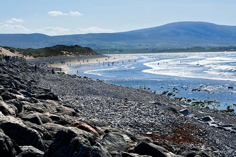 Strandhill Beach in Sligo with its pebbled shoreline, surfers in the water, and distant green hills.