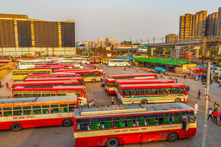 Um terminal de ônibus movimentado com vários ônibus vermelhos estacionados e pessoas andando, cercado por edifícios altos e infraestrutura.