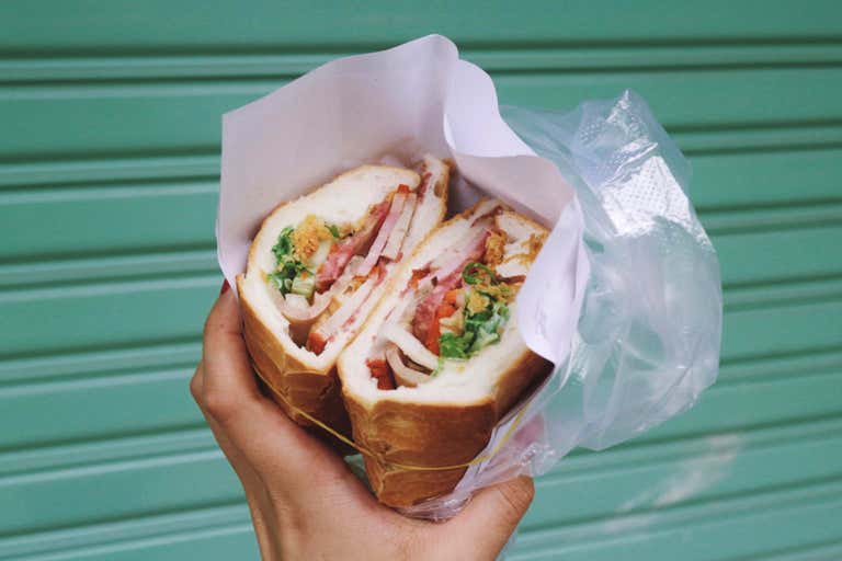 A hand holding a sandwich wrapped in paper against a teal background