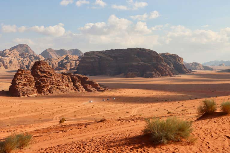 Vast desert landscape of Wadi Rum with orange sand, rock formations, and distant mountains under blue sky.