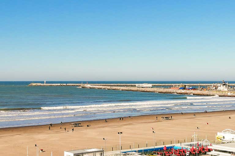 Uma praia de areia com algumas pessoas caminhando ao longo da costa, com um píer e edifícios ao fundo, sob um céu azul claro.