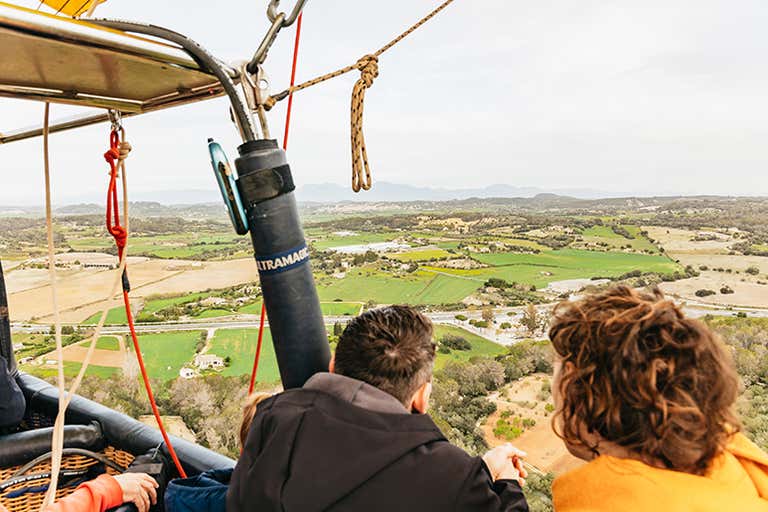 A couple enjoys a hot air balloon ride over the scenic landscape of Mallorca, with fields and mountains in the distance.