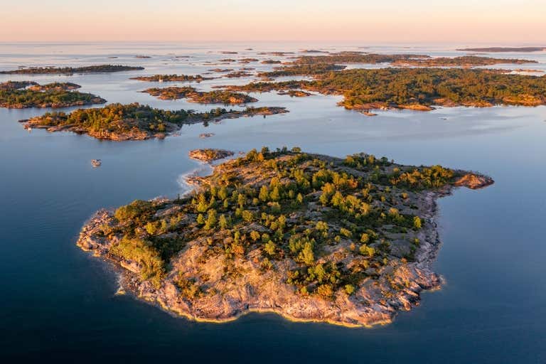 Vista aérea do arquipélago de Estocolmo ao pôr do sol, com várias pequenas ilhas arborizadas espalhadas em águas azuis calmas.