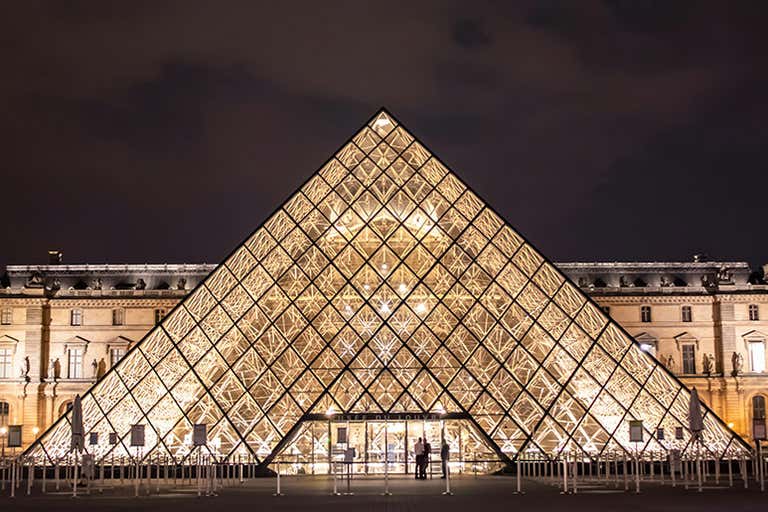 The illuminated Louvre Pyramid at night with historic museum buildings in the background.