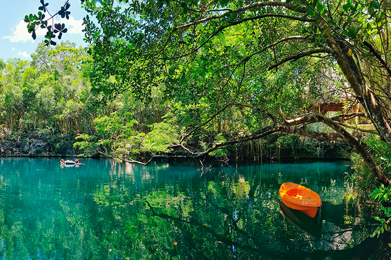 An orange kayak floats on a tranquil, green cenote surrounded by lush trees, with swimmers enjoying the serene water.