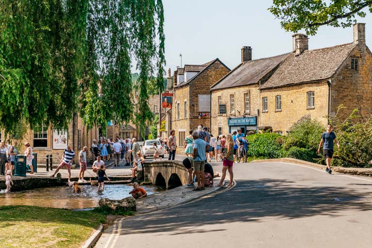 A bustling street scene in Bourton-on-the-Water with people enjoying the sunny day near a small river and stone bridge.