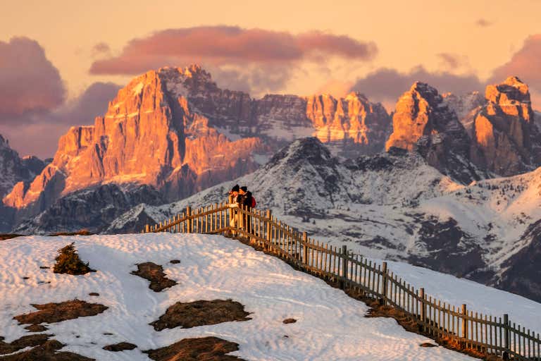 Ein schneebedeckter Aussichtspunkt mit Holzzaun und Blick auf die Dolomiten, die bei Sonnenuntergang orange leuchten - ein einmaliger Anblick