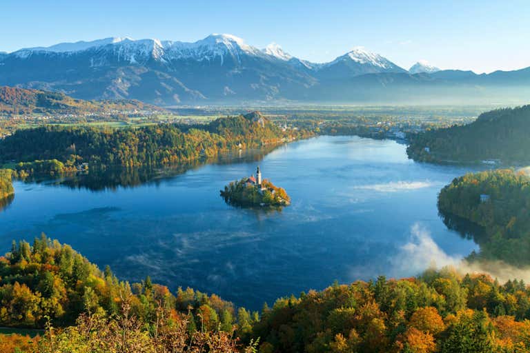 Sunrise over Lake Bled with its iconic island church, surrounded by autumn forests and snow-capped Julian Alps in the background
