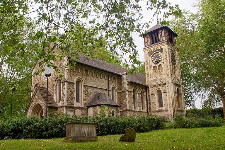 St. Pancras Old Church surrounded by trees and gravestones with distinctive clock tower and stone facade in Camden