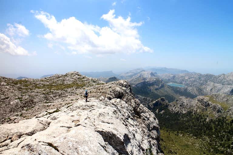 A hiker walking along a rocky ridge with a panoramic view of mountains and a lake.