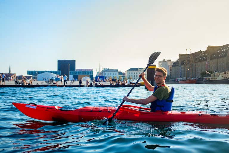A person kayaking in a red kayak on a sunny day in Copenhagen harbor.