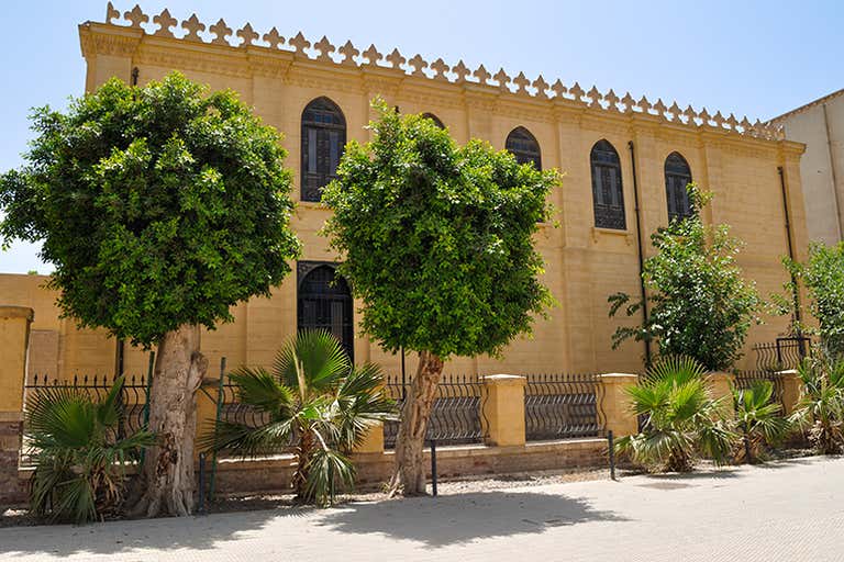 Exterior of Ben Ezra Synagogue in Cairo with arched windows, decorative roofline, and trees lining the foreground.