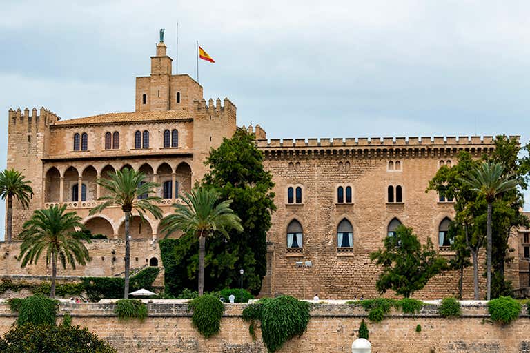 A historic castle with a Spanish flag on top, surrounded by palm trees.