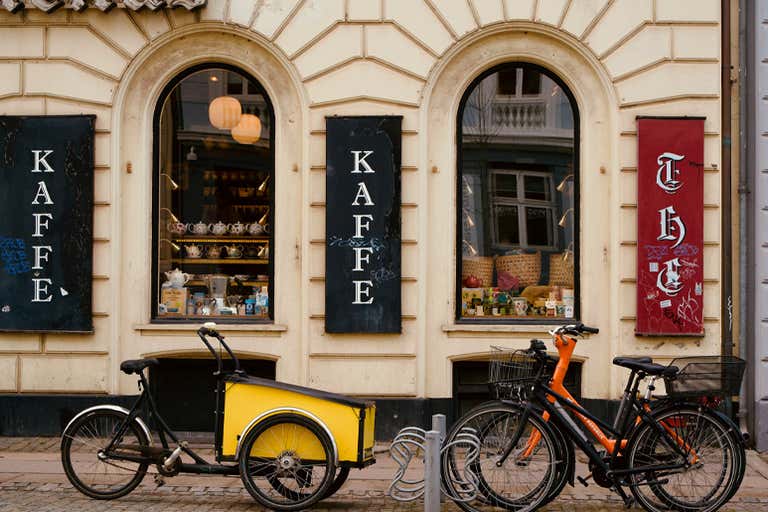 A cozy café storefront with "Kaffe" signs, arched windows displaying teapots, and bicycles parked outside.