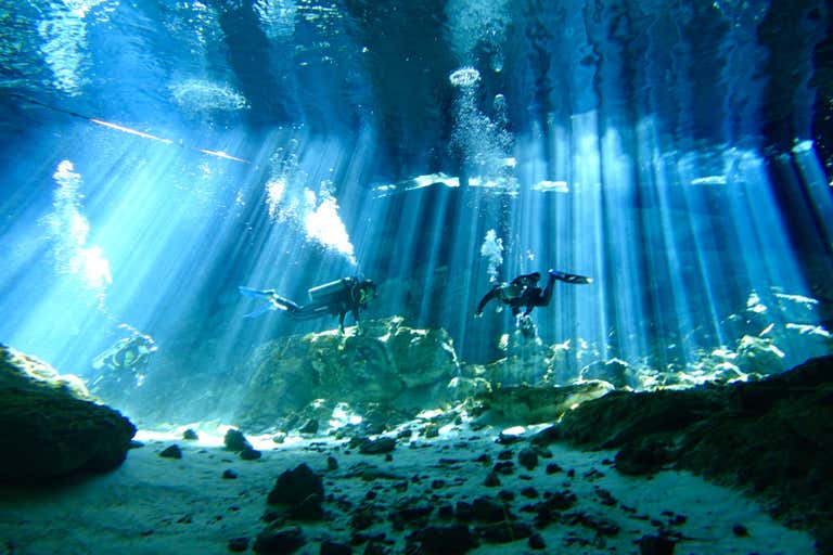 Scuba divers explore an underwater cave illuminated by dramatic beams of sunlight filtering through the crystal-clear water.
