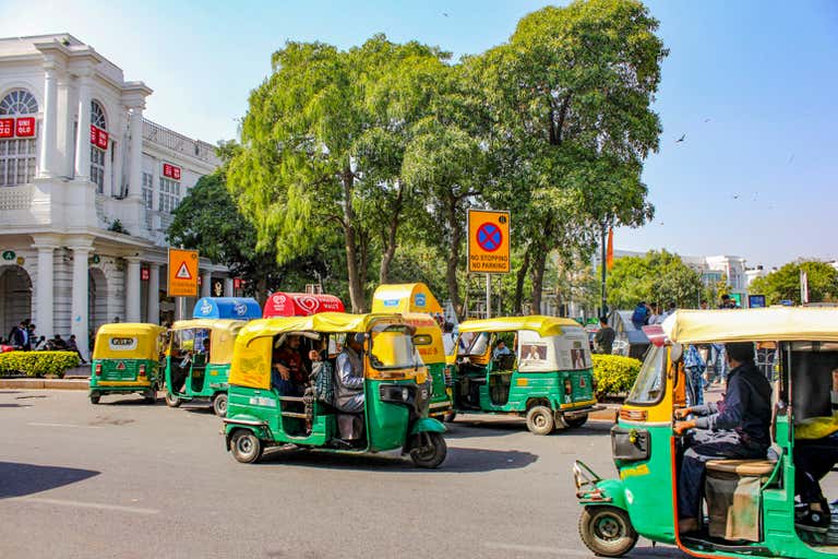 Auto rickshaws em Connaught Place, Nova Délhi, com árvores e edifícios.