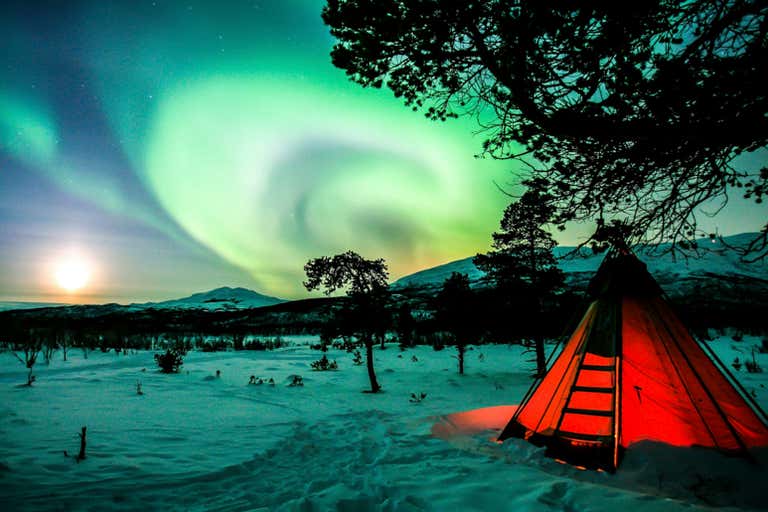 A aurora boreal gira em torno de uma tenda vermelha iluminada em uma paisagem nevada da Lapônia sueca.