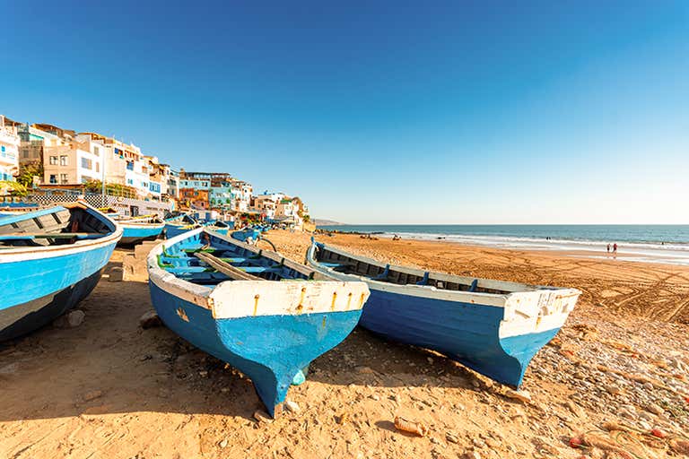 Barcos de pesca azules alineados en la playa de arena de Agadir, con vistas a los edificios y al cielo azul detrás.