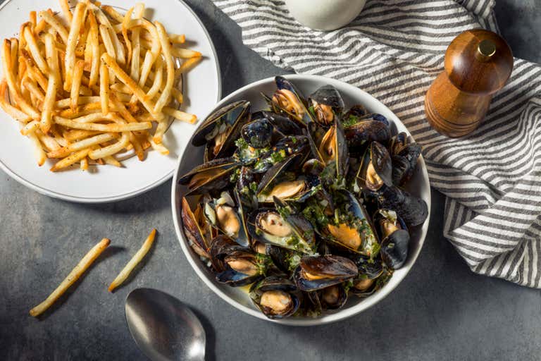 A bowl of cooked mussels with herbs next to a plate of French fries on a striped cloth.