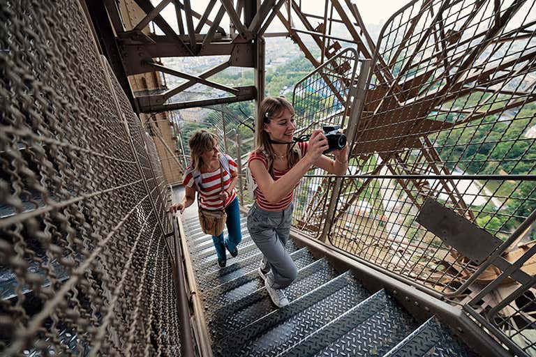 Dois visitantes sobem as escadas da Torre Eiffel, capturando a vista de Paris em junho. Vigas de aço e vegetação visíveis ao fundo.