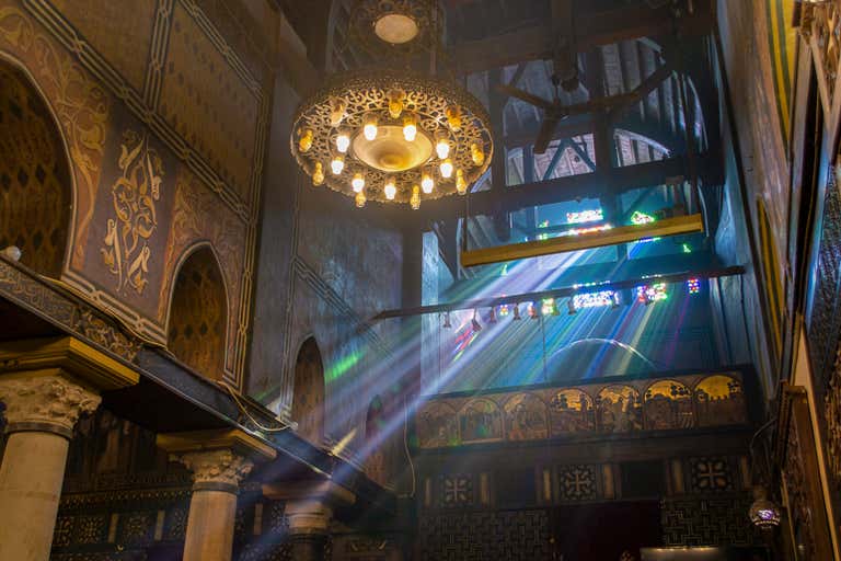 Sunlight streams through stained-glass windows, illuminating ornate carvings and a grand chandelier inside a historic church.