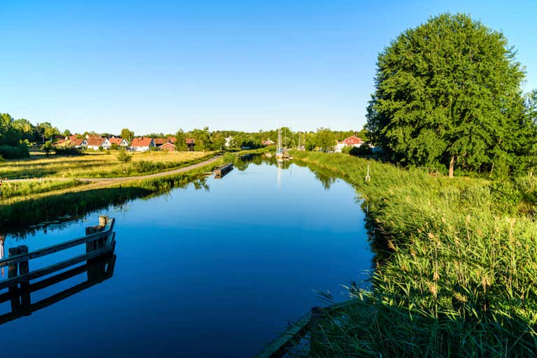 Canal tranquilo na zona rural da Suécia com reflexo de árvores e casas de telhado vermelho em águas paradas.