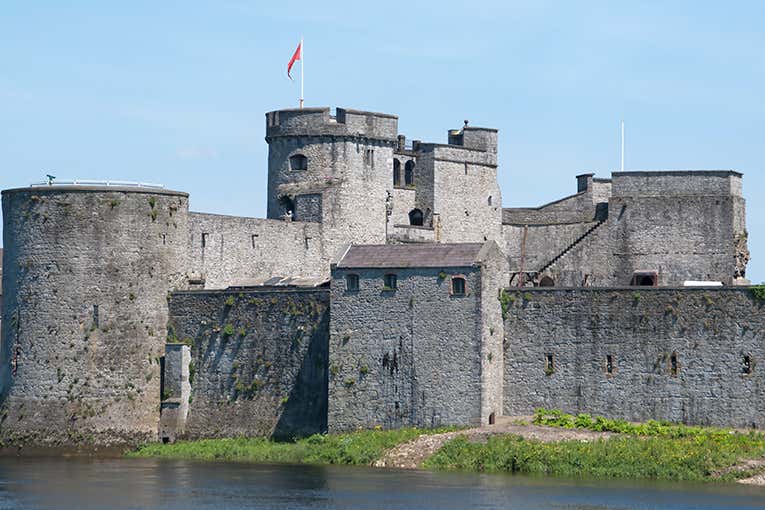 A historic stone castle with a river in the foreground and a flag on top.