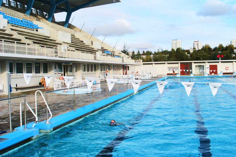 Una piscina al aire libre con carriles y algunas personas nadando, rodeada por una estructura similar a un estadio y árboles al fondo.