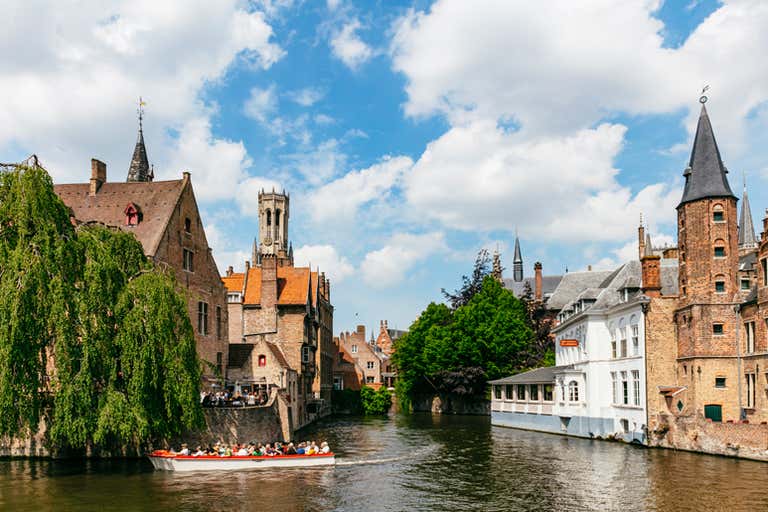 Boat tour on a canal in Bruges with historic brick buildings and lush greenery under a partly cloudy sky.