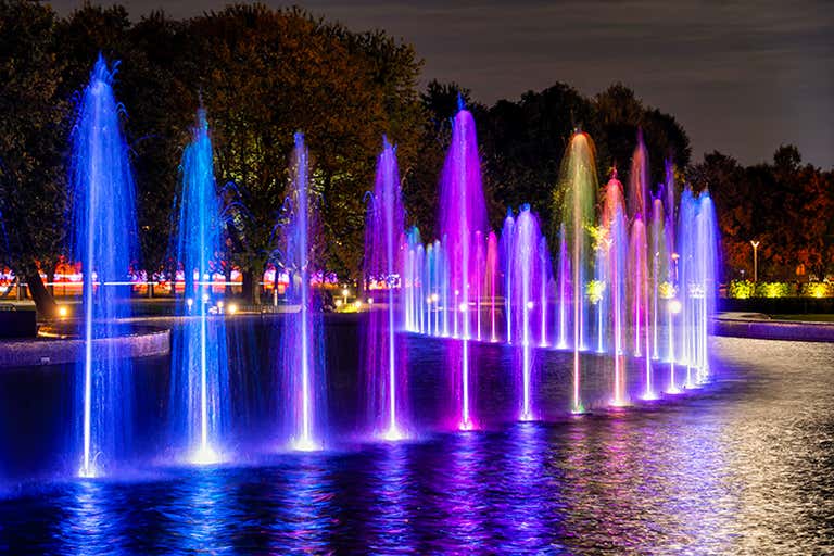 Fontes coloridas e iluminadas em um parque à noite, com jatos de água em tons de azul, roxo e rosa.