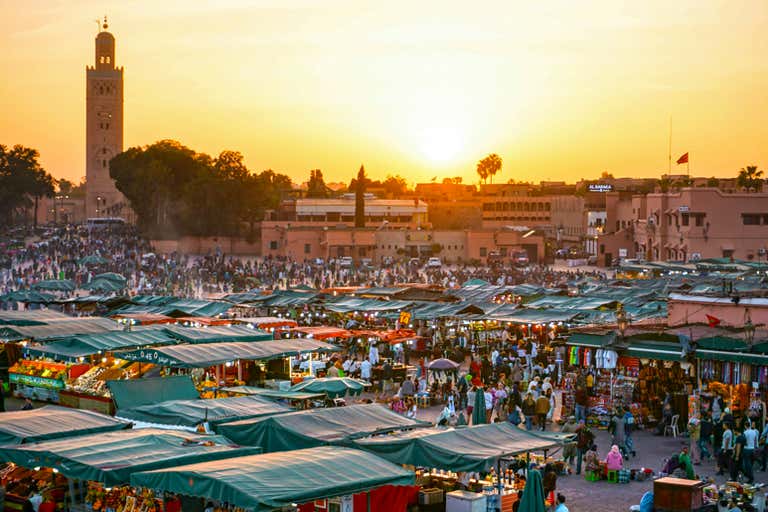 Una bulliciosa plaza del mercado de Marrakech al atardecer con la torre de una mezquita al fondo.