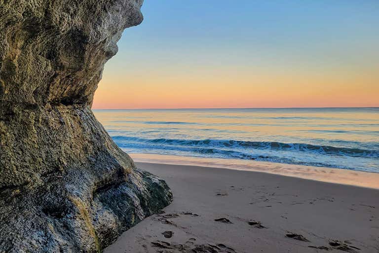 Pôr do sol em uma praia tranquila com ondas suaves, pegadas na areia e um penhasco rochoso à esquerda sob um céu em tons pastéis.