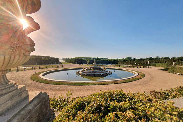 Sunny view of Versailles’ gardens with a fountain, circular pond, and decorative urn in the foreground.
