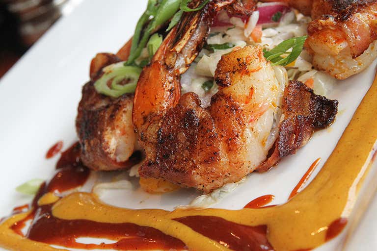 Bacon-wrapped grilled shrimp served on a bed of slaw with drizzles of spicy mustard and barbecue sauce on a white plate.