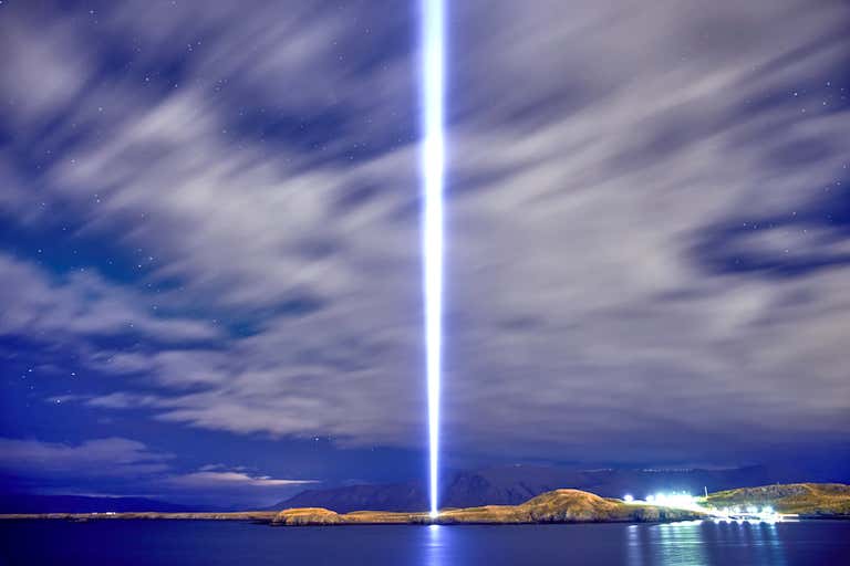 Un haz de luz brillante se extiende hacia el cielo desde una zona costera bajo un cielo nocturno nublado.