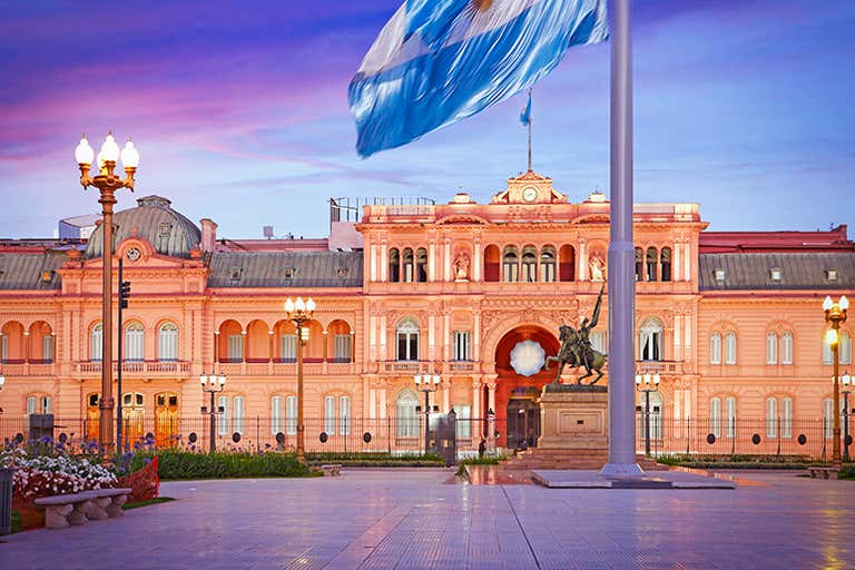 Casa Rosada presidential palace in Buenos Aires with Argentine flag and monument at sunset.