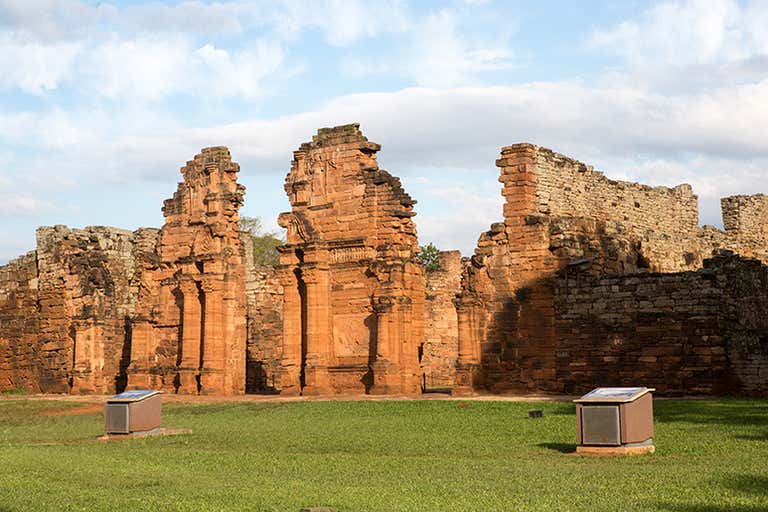 Ruínas de San Ignacio Miní com paredes de pedra vermelha e grama verde.