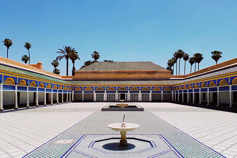 Un patio con una fuente en el centro, rodeado de arcos y palmeras bajo un cielo azul despejado.