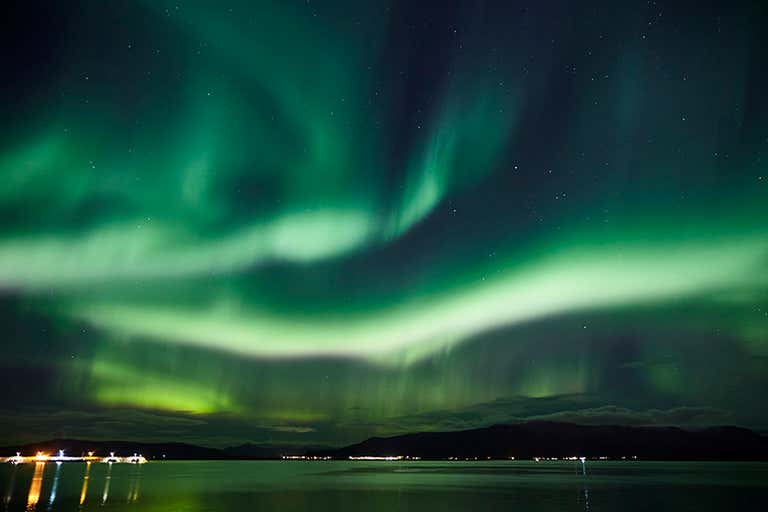 Aurora boreal sobre una masa de agua en calma con luces lejanas en el horizonte.