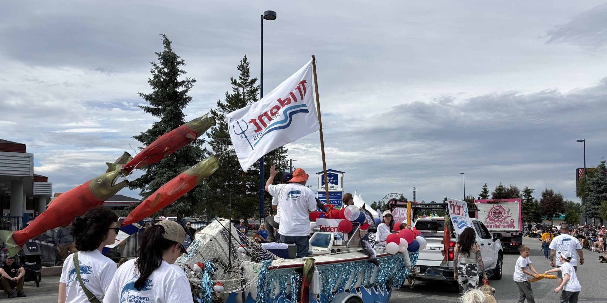 Parade float decorated with oversized salmon, fishing nets, and red, white, and blue balloons travels down a city street. A white Trident Seafoods flag waves above the float as spectators line the route under a cloudy sky.
