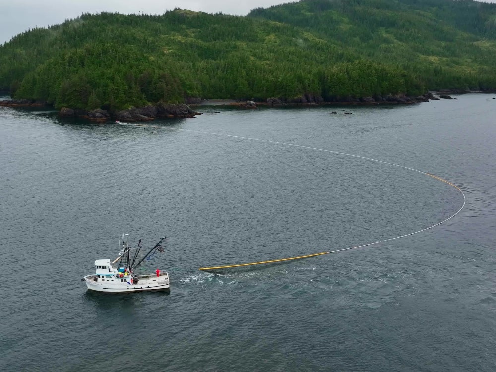Aerial view of a commercial fishing boat setting a seine net in calm Alaska waters, with a tree-covered coastline and rugged shoreline in the background.