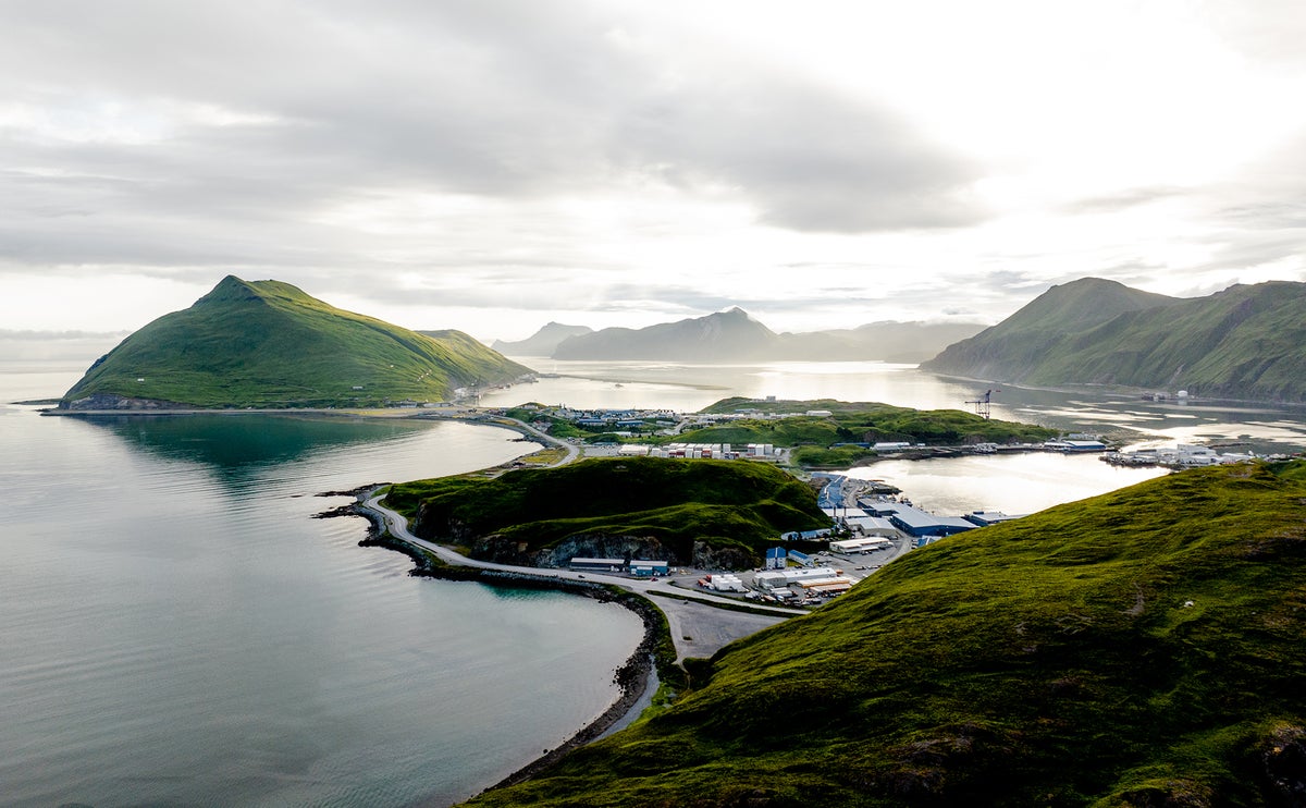 Wide aerial landscape of Dutch Harbor, Alaska, an active fishing and seafood processing hub in the Aleutian Islands, surrounded by mountainous coastline and ocean waters.