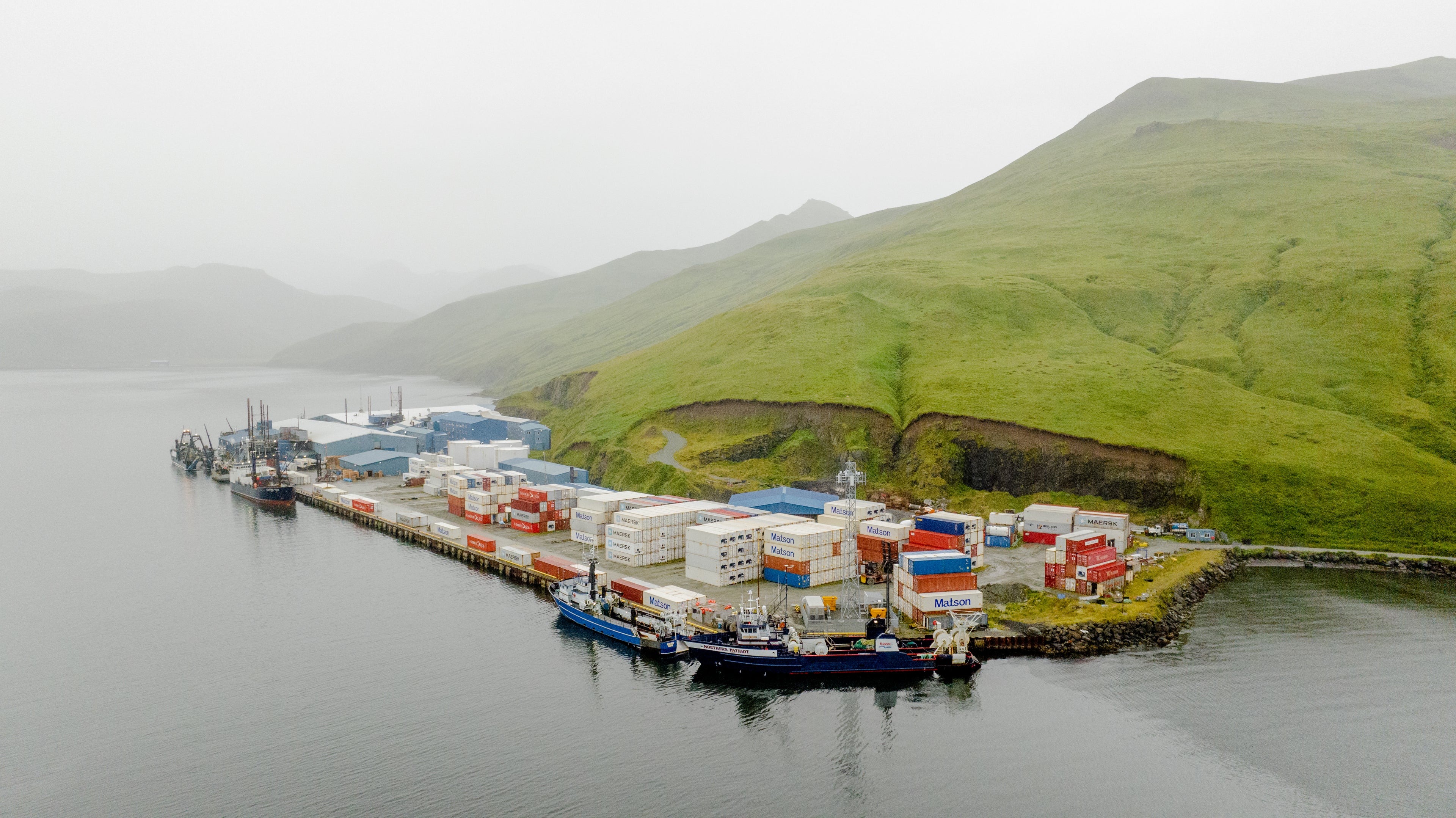 Container ports with docked fishing vessels are nestled among green hills under an overcast sky in Dutch Harbor, Alaska. 