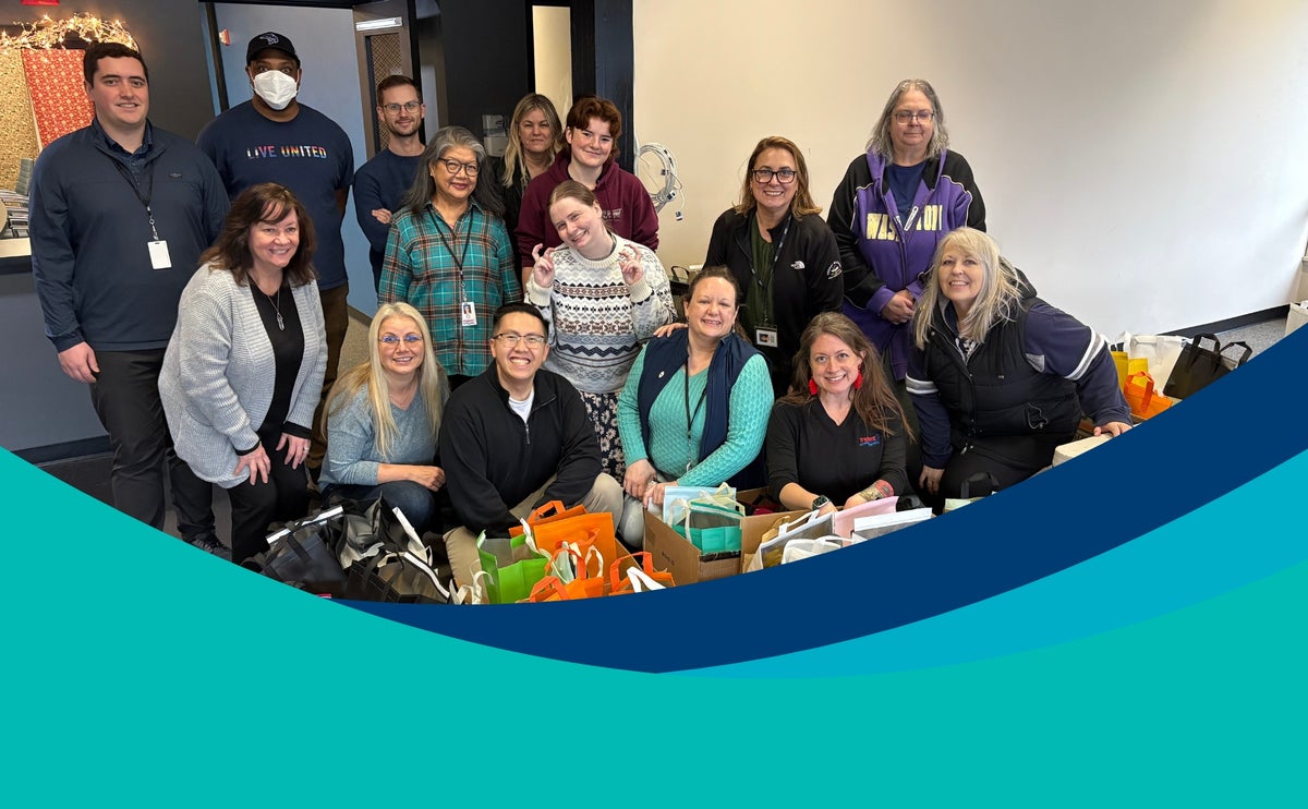 Trident Seafoods employees pose together indoors, gathered behind rows of colorful gift bags and donation items, with teal and navy graphic shapes overlaid along the bottom of the image.