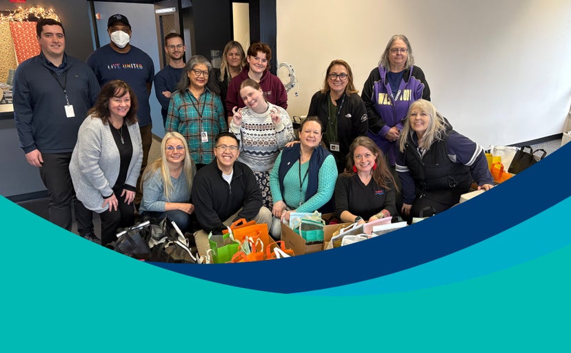 Trident Seafoods employees pose together indoors, gathered behind rows of colorful gift bags and donation items, with teal and navy graphic shapes overlaid along the bottom of the image.