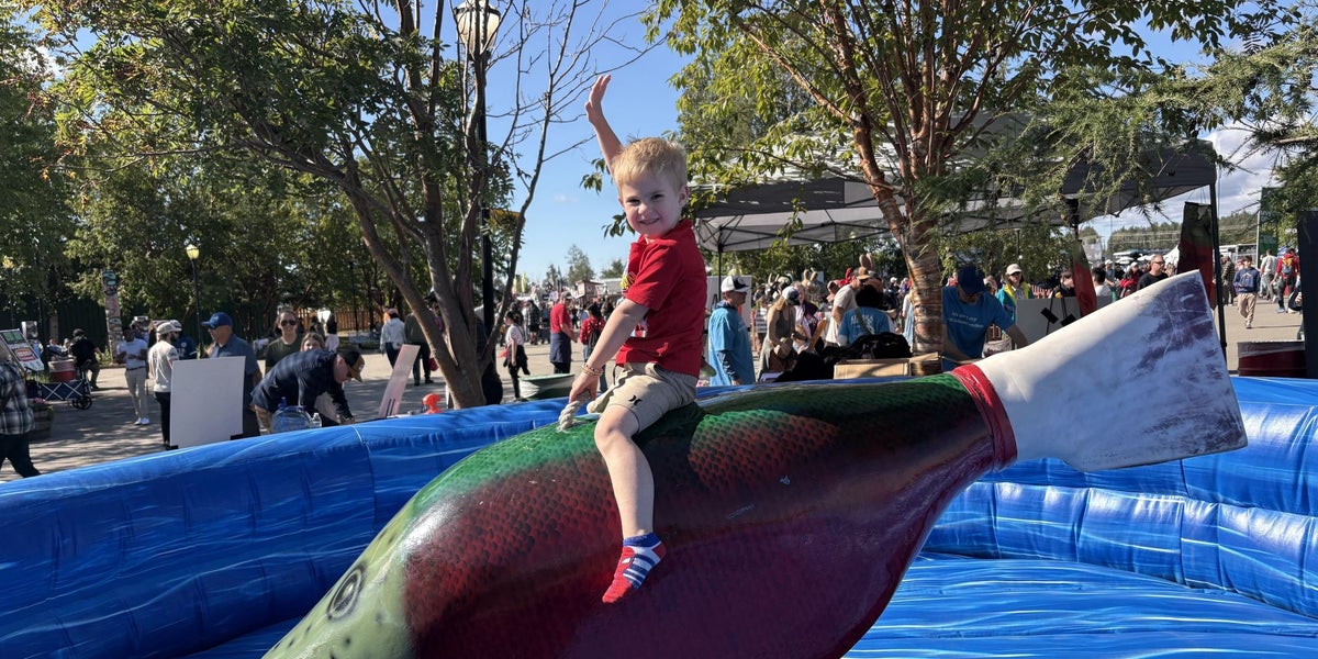 Child sitting on a large fish-shaped rodeo attraction inside an inflatable blue arena at a community festival, with trees, vendor tents, and crowds visible in the background on a sunny day.
